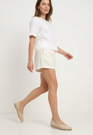 Kobieta idąca ubrana w biały krótki top, białe spodenki i beżowe płaskie buty na jasnym tle.