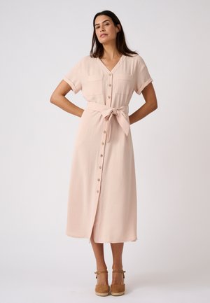 Mujer con cabello largo y oscuro, vestida con un vestido midi rosa claro abotonado, con bolsillos y cinturón en la cintura, lleva tacones marrones con correa en el tobillo, de pie con las manos en las caderas.