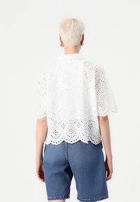 Witte geborduurde shirt met korte mouwen, met een scalloped zoom en een gaatjespatroon, gecombineerd met blauwe denim shorts. Het ontwerp heeft een kraag.