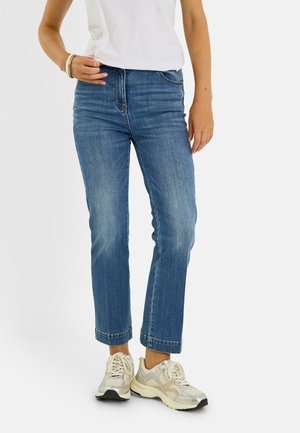 Personne portant un jean bleu taille moyenne, un t-shirt blanc et des baskets blanches avec des accents dorés, debout devant un fond clair uni.