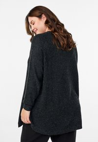 Prevelik črn pulover iz teksturirane tkanine, z okroglim izrezom in daljšim hrbtnim robom, dolgimi rokavi in sproščenim krojem.