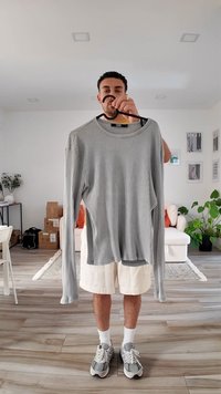 Camisola cinza de manga longa com gola redonda, pendurada numa cabide, apresentando aberturas laterais e um corte descontraído.