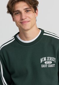 Felpa verde scuro con strisce bianche sulle maniche, dotata di un logo stampato "New Jersey East Coast dal 1985" sul petto.