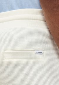 Κοντινή λήψη λευκού υφασμάτινου παντελονιού με πίσω τσέπη που φέρει μια μικρή ορθογώνια ετικέτα "Lindbergh", που φοριέται από άτομο με μέτρια σκούρο δέρμα.