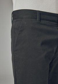 Schwarze Baumwollhose mit flachem Bauch, seitlichen Taschen und einem mittelhohen Taillenbund. Der Stoff wirkt glatt mit einem leichten Glanz.
