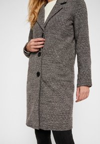 Šedý texturovaný kabát s přiléhavým střihem, jednoradovým designem, klopeným límcem, třemi černými knoflíky a bočními kapsami.