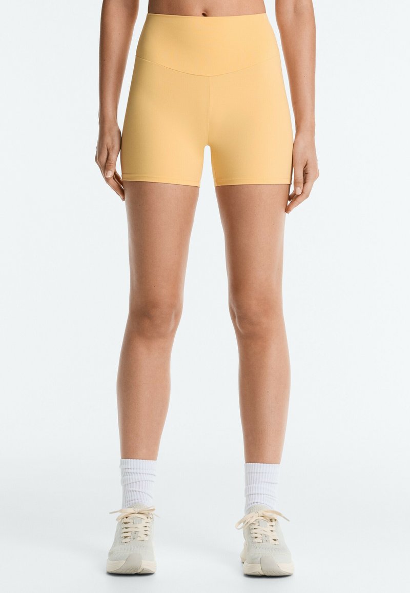 Femme portant un short cycliste jaune taille haute avec des chaussettes blanches et des baskets beiges, debout devant un fond clair uni.