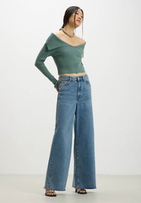 Maglione a coste verde chiaro con spalle scoperte abbinato a jeans in denim blu a vita alta e fit abbondante. La modella indossa sandali neri con sabot.
