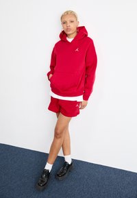 Rød hettegenser med lomme foran og hvit logo, kombinert med matchende røde shorts. Svart plattform sko og hvite sokker. Enkel bakgrunn.