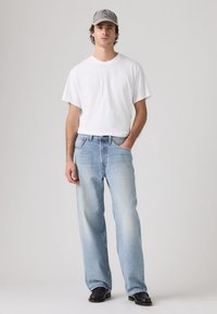 Νέος άνδρας που φοράει γκρι καπέλο, λευκό μπλουζάκι, ανοιχτό μπλε τζιν και μαύρα παπούτσια, στέκεται με το ένα χέρι στην τσέπη του μπροστά σε ένα απλό φόντο.