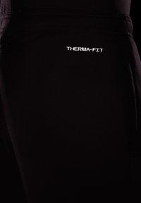 Czarne spodnie Therma-Fit o gładkiej teksturze. Posiadają pionowe, białe logo "THERMA-FIT" na lewym udzie. Smukły krój oraz funkcjonalne kieszenie.