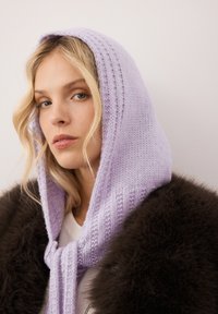 Jonge vrouw met blond haar, die een lila gebreide sjaal met kap en een donkerbruine bontjas draagt, en lichtjes opzij kijkt.