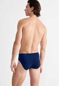Mann mit schulterlangen Haaren, der dunkelblaue Unterwäsche trägt und mit dem Rücken zur Kamera vor einem schlichten weißen Hintergrund steht.