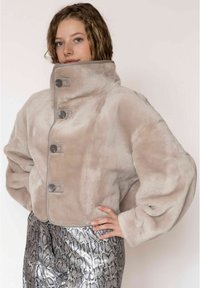 Korte, zachte beige jas met een hoge kraag en oversized mouwen; heeft vier donkere knopen aan de voorkant en een zachte textuur.