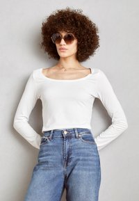 Camisa blanca de manga larga con escote cuadrado, combinada con vaqueros de mezclilla azul de cintura alta. La modelo lleva gafas de sol marrones y un collar minimalista.