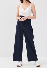 Femme en haut sans manches blanc, pantalon large navy avec ceinture et baskets blanches, se tenant devant un fond clair.