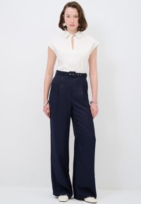 Haut blanc à manches courtes avec un col à trou, associé à un pantalon large taille haute bleu marine et des chaussures de couleur claire. Accessoires simples.