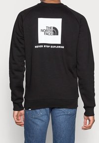 Svart sweatshirt gjord av bomullsblandning, med en fyrkantig vit logotyp på baksidan och texten "NEVER STOP EXPLORING" under.