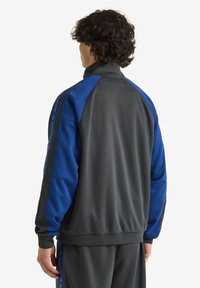 Giacca sportiva grigia e blu, con zip completa e colletto alto, polsini a coste e pannelli laterali. Il tessuto sembra liscio con una leggera lucentezza.