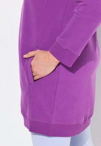 Lila Fleece-Tunika mit langen Ärmeln, seitlichen Taschen, gerippten Bündchen und lockerer Passform. Glatte Textur, leichter Stoff.