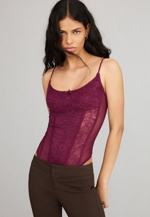 Jonge vrouw met lang donker haar, gekleed in een paarse kanten top en een bruine broek tegen een effen grijze achtergrond.
