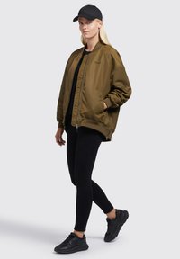 Olivovo zelená bomber jacket s predným zipsom, ribovanými manžetami a bočnými vreckami, v kombinácii s čiernymi legínami a čiernymi teniskami. Na hlave čierna čiapka.