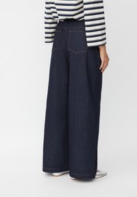 Pantalones de mezclilla oscura de pierna ancha con bolsillos traseros y costuras naranjas en contraste. Combinados con una blusa de manga larga a rayas en navy y blanco.