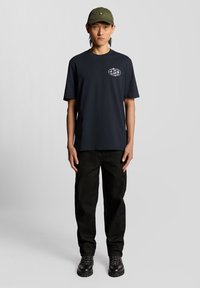 Tamsiai mėlynas medvilninis marškinėlis su baltu grafiniu logotipu kairėje krūtinės pusėje, derinamas su juodomis tekstūruotomis kelnėmis ir juodais raišteliais suvarstytais batais; žalia kepurė.