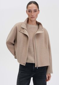 Beige wollen jas met een hoge kraag, rits aan de voorkant en drukknoopsluitingen, gelaagd over een lichtbeige trui, gecombineerd met zwarte jeans.