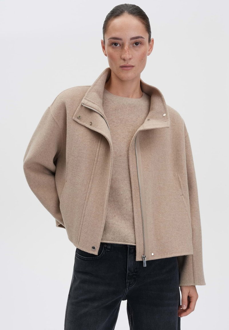 Beige wollen jas met een hoge kraag, rits aan de voorkant en drukknoopsluitingen, gelaagd over een lichtbeige trui, gecombineerd met zwarte jeans.