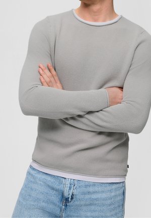 Mann trägt einen hellgrauen strukturierten Pullover mit langen Ärmeln und hellblauen Jeans, die Arme vor der Brust verschränkt, stehend vor einem schlichten Hintergrund.