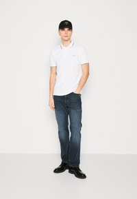 Witte polo met zwarte afwerking, blauwe jeans en zwarte schoenen. Het model draagt een zwarte pet met witte tekst en staat tegen een neutrale achtergrond.