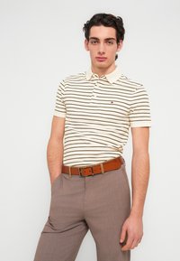 Πόλο μπλούζα με κοντά μανίκια σε κρεμ απόχρωση με λεπτές μαύρες ρίγες, διακοσμημένη με γιακά και μικρό λογότυπο, συνδυασμένη με ανοιχτό καφέ παντελόνι φόρμας και ζώνη.