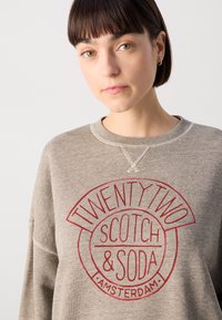 Sivá mikina s okrúhlym červeným grafickým potlačou s nápisom "TWENTY TWO SCOTCH & SODA AMSTERDAM." Obsahuje rebrovaný výstrih a šité detaily.
