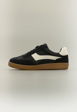 Zwarte lage sneaker met witte zijstreep en hielstuk, bruine rubberen zool en vetersluiting.
