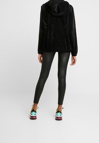 Zwarte corduroyjas met een capuchon, gecombineerd met glanzende zwarte leggings en kleurrijke sneakers met een speelse print en een aflopende zool.