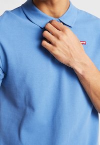 Polo bleu clair en tissu texturé, avec un col classique et un petit logo Levi's rouge près du col.