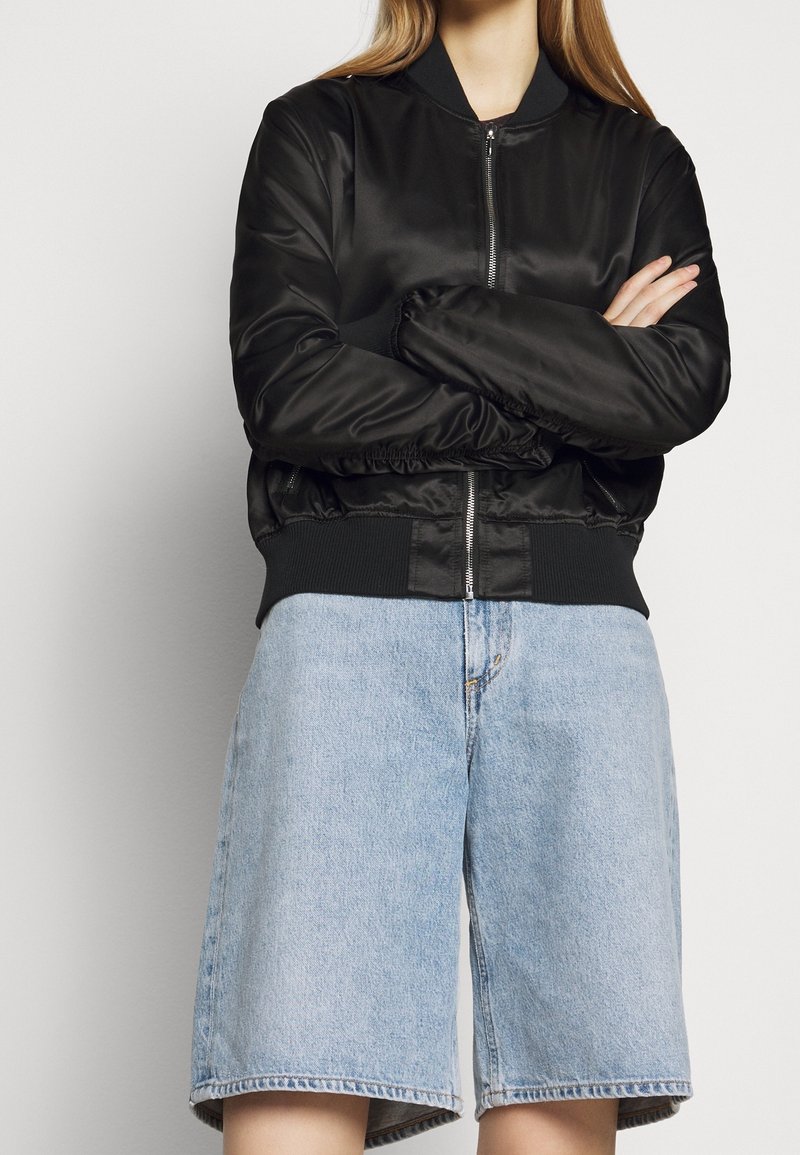 Personne portant une veste aviateur noire à fermeture éclair avec poignets et ourlet côtelés, associée à un short en denim bleu clair taille haute.