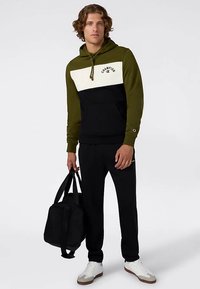 Felpa con cappuccio con maniche verdi, pannello bianco sul petto e corpo nero; abbinata a pantaloni neri e scarpe bianche, con una borsa nera in mano.