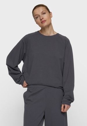 Kvinde med lys hud og blondt hår iført en løs mørkegrå sweatshirt og matchende bukser, stående med den ene hånd på hoften på hvid baggrund.