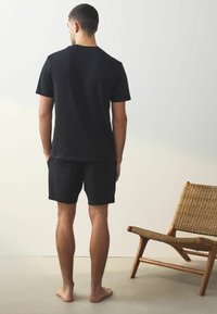 Tricou negru cu mânecă scurtă și pantaloni scurți negri, realizate dintr-un material moale, cu o croială confortabilă. Picioare goale și un fotoliu din ratan în fundal.