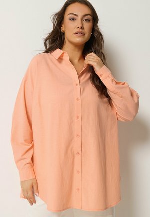 Frau mit langen Haaren, die ein übergroßes, pfirsichfarbenes Hemd trägt und mit einer Hand nahe dem Kragen vor hellem, einfarbigem Hintergrund posiert.