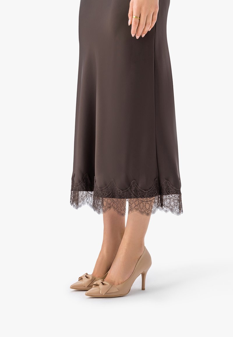 Žena u smeđoj midi suknji s čipkastim obrubom i bež visokim potpeticama s mašnom, stoji s rukama opuštenima uz tijelo.