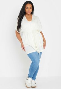 Vit stickad cardigan med knytbälte, korta ärmar och en lös passform, kombinerad med blå smala jeans och vita sneakers med svarta ränder.