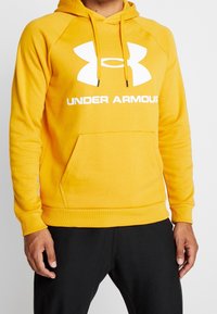 Sudadera amarilla con un bolsillo tipo canguro y puños acanalados. Presenta un gran logotipo blanco de Under Armour en la parte frontal. Material suave de mezcla de algodón.