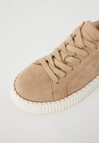 Beige suede sneaker with flat laces, round toe, and a textured rubber sole featuring a rope accent around the edge.