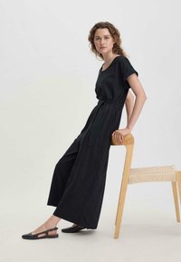 Jumpsuit negro de pierna ancha con mangas cortas y cintura ceñida con cinturón. La modelo está de pie al lado de una silla de madera. Sandalias planas negras con detalles recortados.
