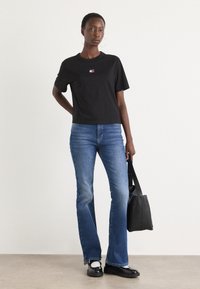 Tricou din bumbac negru cu guler rotund și un logo mic, asortat cu blugi albaști flared și o geantă neagră, completat cu pantofi negri jos.
