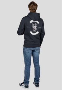 Sudadera negra con un diseño gráfico que presenta una muñeca de tela cosida en la parte posterior, combinada con jeans de un azul claro y zapatillas en blanco y negro.