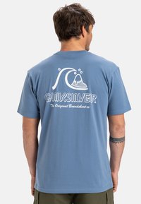 Blauwe katoenen T-shirt met korte mouwen, met een witte golfafbeelding en "Quiksilver" tekst op de achterkant. Rechte pasvorm met geribde halslijn.